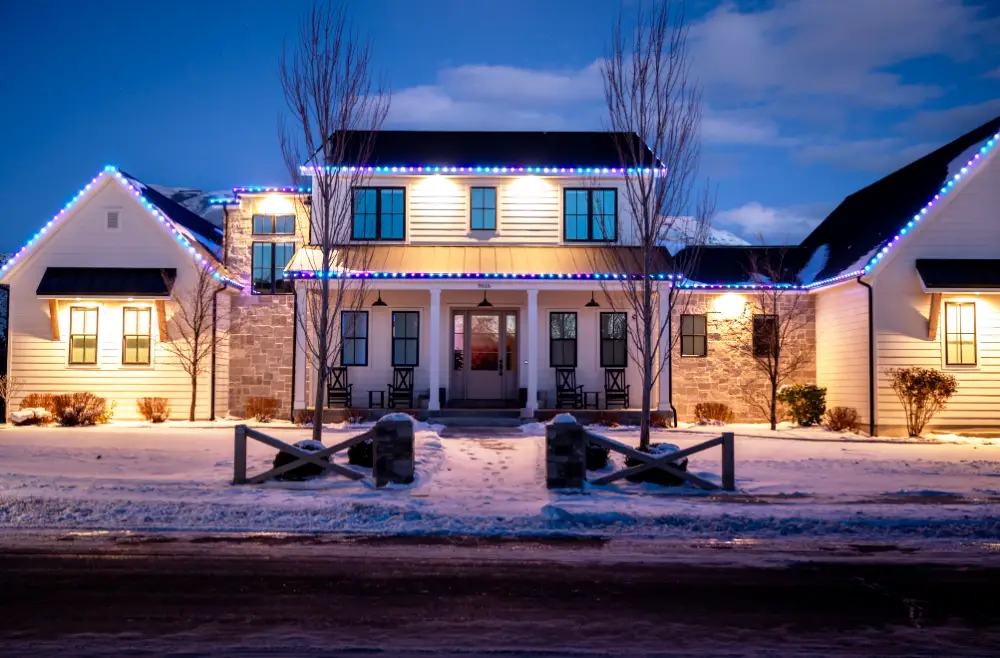White farmhouse-style Utah County home at dusk in winter with Canyon Clear permanent LED lights in blue and purple along the rooflines